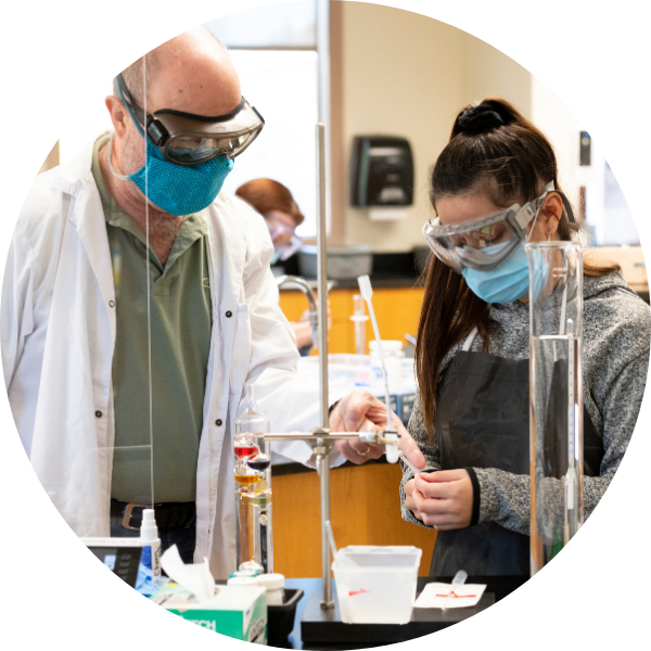 An instructor and a student perform an experiment together in a science lab. Both are wearing surgical masks. 