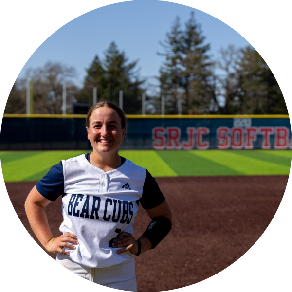 A softball player standing in front of a softball field, hands to her side, in uniform, smiling. 