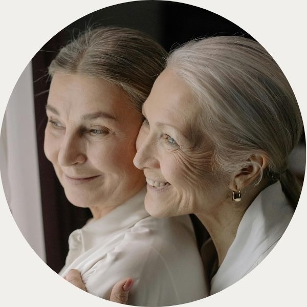 Two older women looking lovingly off into the distance together, smiling. One woman appears to be holding the other woman from behind. 